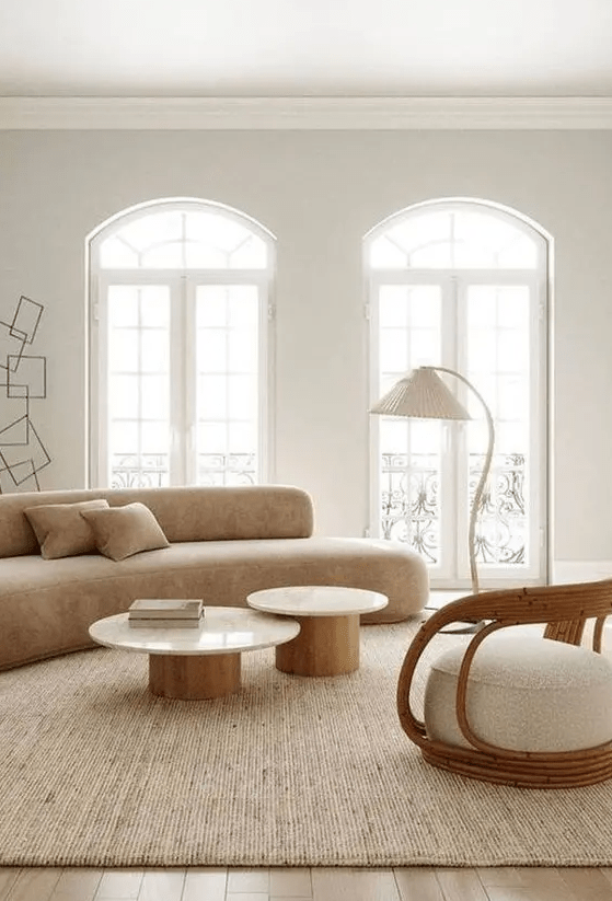 a chic neutral living room with a curved sofa, a duo of coffee tables, a rattan chair, a floor lamp and arched windows