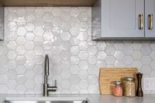 a grey kitchen with white stone countertops and a glossy white hexagon tile backsplash plus stained shelves