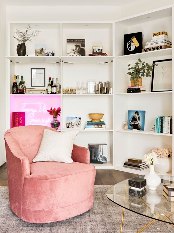 A pretty nook with storage unit and built in lights, a neon sign, a pink curved chair and a glass coffee table