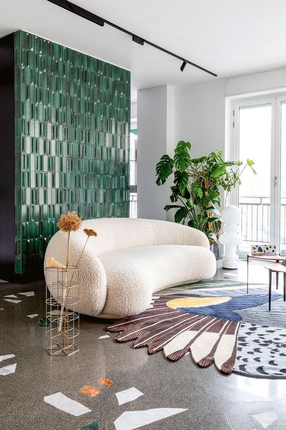 a quirky living room with a green tile wall, a white boucle curved sofa, a colorful rug, side tables and greenery