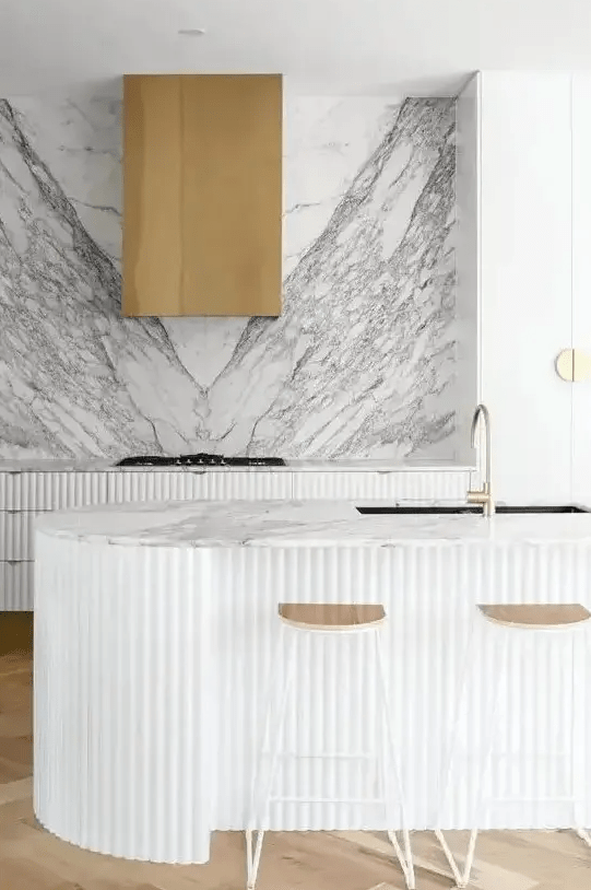 a refined contemporary kitchen with ridged cabinets and a matching curved kitchen island, a white marble backsplash plus a brass hood