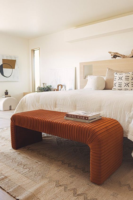 A serene bedroom with a bed and neutral bedding, a rust colored curved bench, a neutral rug and a side table