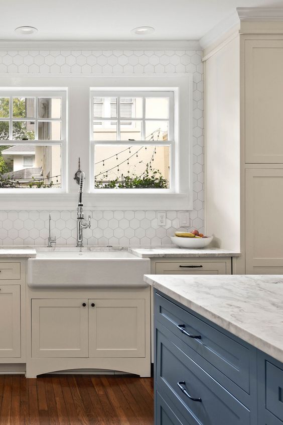a vintage creamy kitchen with white stone countertops, white hexagon tiles on the wall and a blue kitchen island