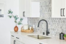 a whire kitchen with shaker cabinets, white stone countertops, a grey hexagon tile backsplash and black fixtures