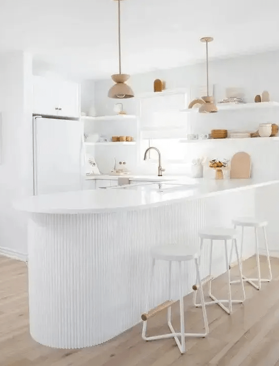 An all white kitchen with sleek plain cabinets, a curved ribbed kitchen island that is an accent here, open shelving instead of upper cabinets
