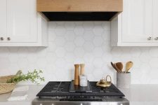 an elegant white kitchen with shaker cabinets, a hood with a wooden hem and a white hexagon tile backsplash plus neutral countertops