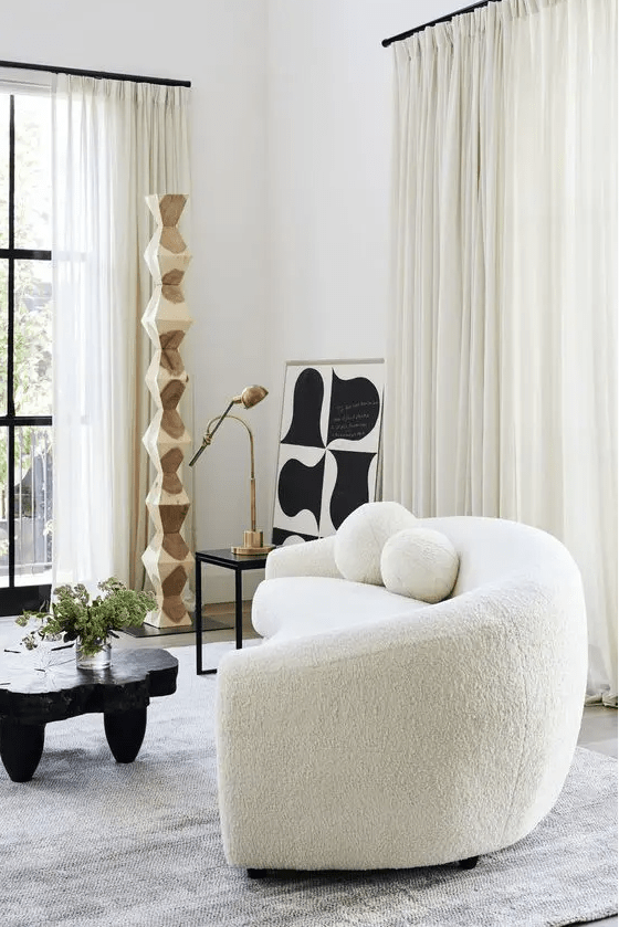 an eye-catchy space with a white boucle curved sofa, a black tree slice table, a side table with a lamp and some abstract art