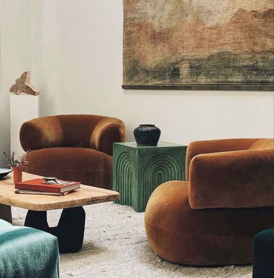 An eye catchy space with rust colored curved chairs, a green stone side table and a coffee table, an oversized artwork and blue poufs