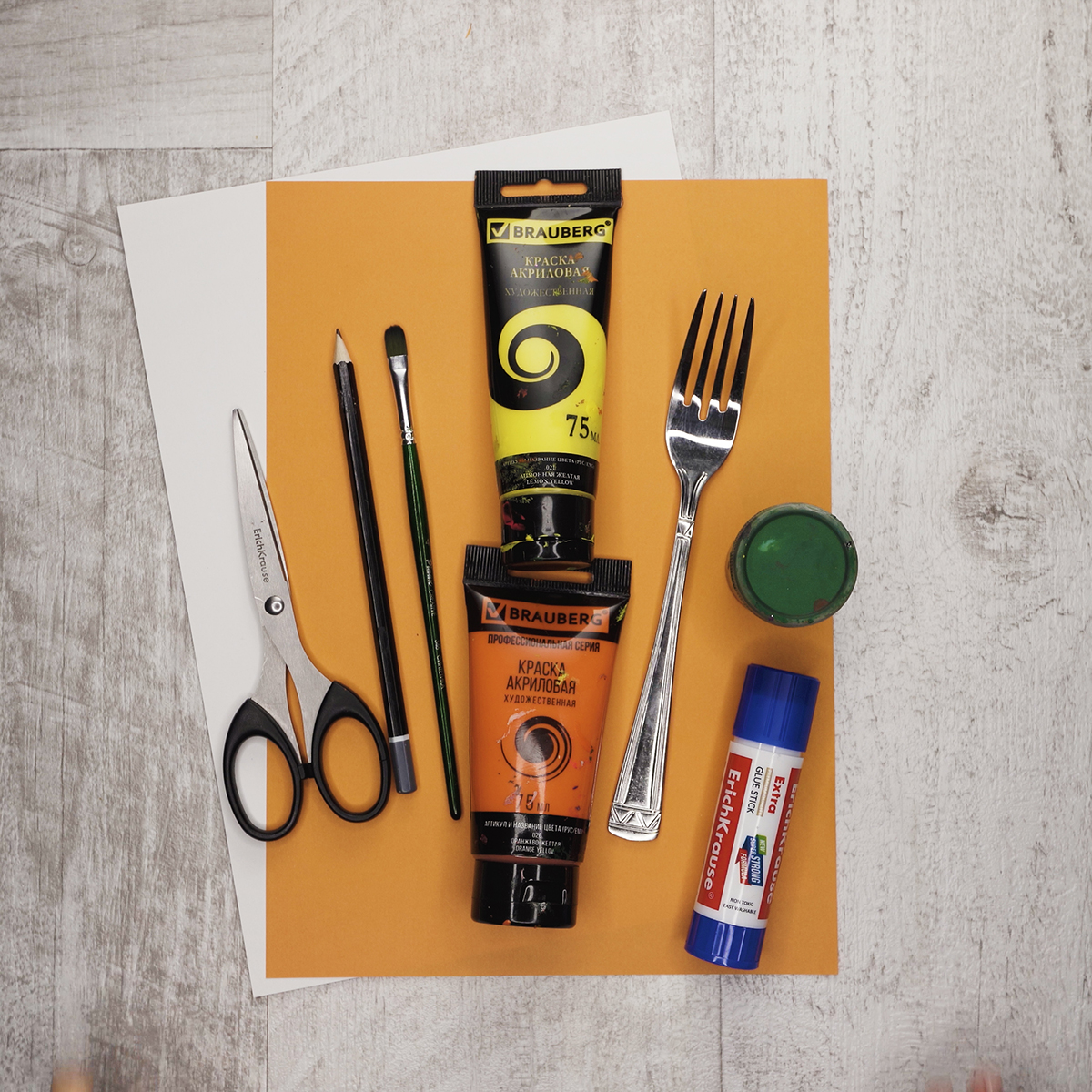 The supplies are orange and white paper, yellow, green and orange paints, a glue stick, a fork, a pencil, a brush and scissors