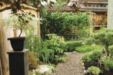a cozy and cool garden with a gravel pathway, greenery and shrubs, a pedestal with a potted plant and some stones and rocks