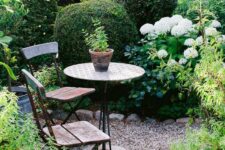 a lush green garden with shrubs, greenery and trees, with white flowers, a small space done with gravel and simple garden furniture