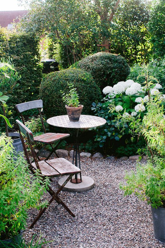a lush green garden with shrubs, greenery and trees, with white flowers, a small space done with gravel and simple garden furniture