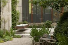 a modern and relaxed garden with a gravel space, wooden chairs and a table, greenery, shrubs and trees and a suspended bench
