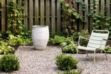 a modern backyard done with pea gravel, with greenery and shrubs, a white chair, a large planter and some stepping stones