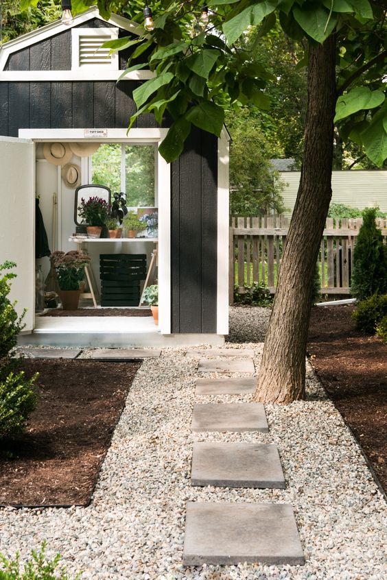 a modern backyard with gravel and concrete stepping stones, a tree and shrubs and a shed for gardening