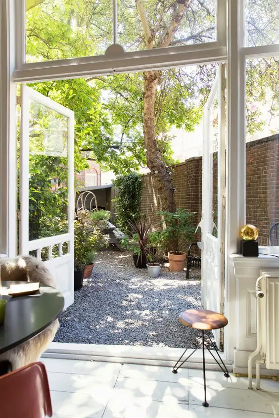 a modern gravel backyard with a tree, potted plants and blooms, some garden furniture is a shadowy and welcoming space