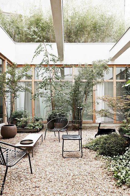 a modern inner yard with gravel on the ground, modern black furniture, a bench, greenery and some trees is chic