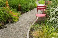 a pretty green garden with greenery and shrubs, some blooms, decor, red chairs and a gravel path with rock edging