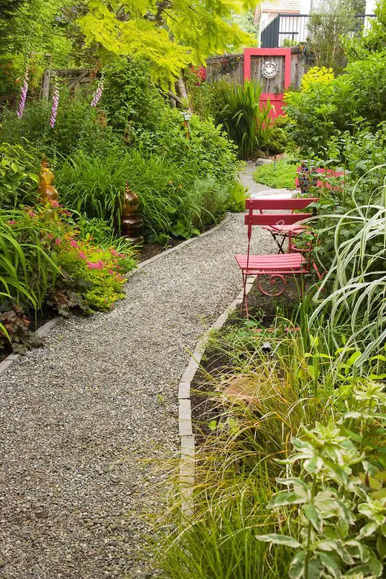 a pretty green garden with greenery and shrubs, some blooms, decor, red chairs and a gravel path with rock edging