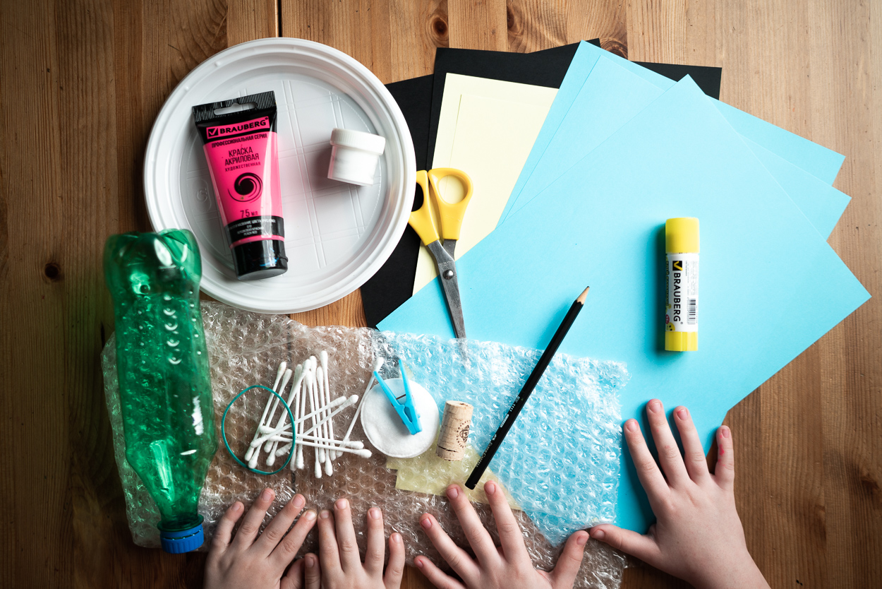 The supplies are colored paper, acrylic and gouache paints, a glue stick, a pencil, scissors, a marker, a rubber elastic, overwrapping, cotton buds, a clothes peg, cotton pegs and wine corks, an empty plastic bottle.