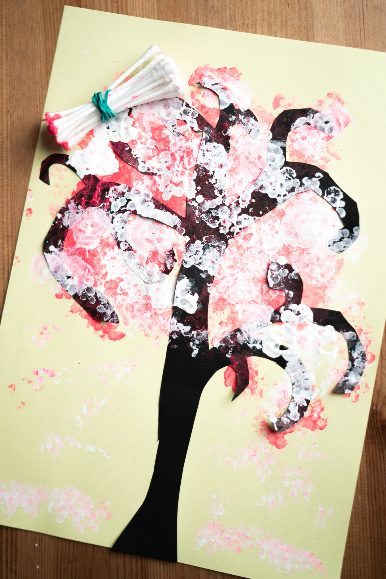 Painting With Cotton Buds
Take a black sheet of paper and draw a tree with a pencil, cut it out. Glue the tree to a blue sheet, take a bunch of cotton buds, secure them with an elastic. Dip the buds into pink paint and stamp them on the tree and all around to imitate blooms. Add some white ‘blooms’, too, using white paint.