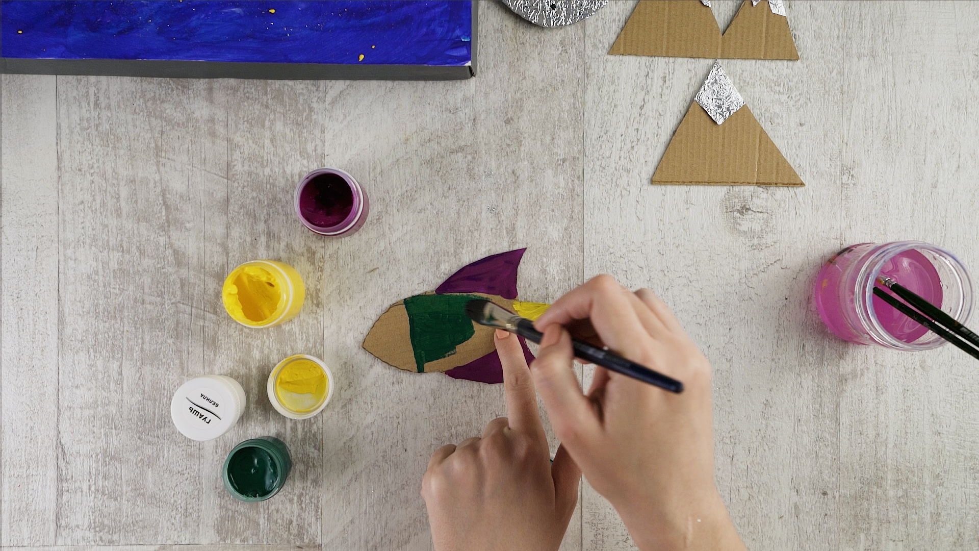 Draw a little rocket on cardboard and then cut it out – your may use the idea from the tutorial or choose your own shape. Paint it in bold colors, here – purple, yellow, white and green, and add some glitter. Glue some cardboard pieces to the rocket.