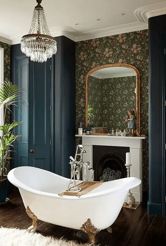 A Victorian style bathroom with moody wallpaper walls, a non working fireplace, a clawfoot bathtub, navy wardrobes, a crystal chandelier and plants