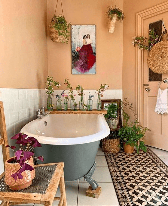 a bold boho meets vintage bathroom with buttermilk walls, a bold rug, a grey clawfoot tub, potted greenery and wicker touches