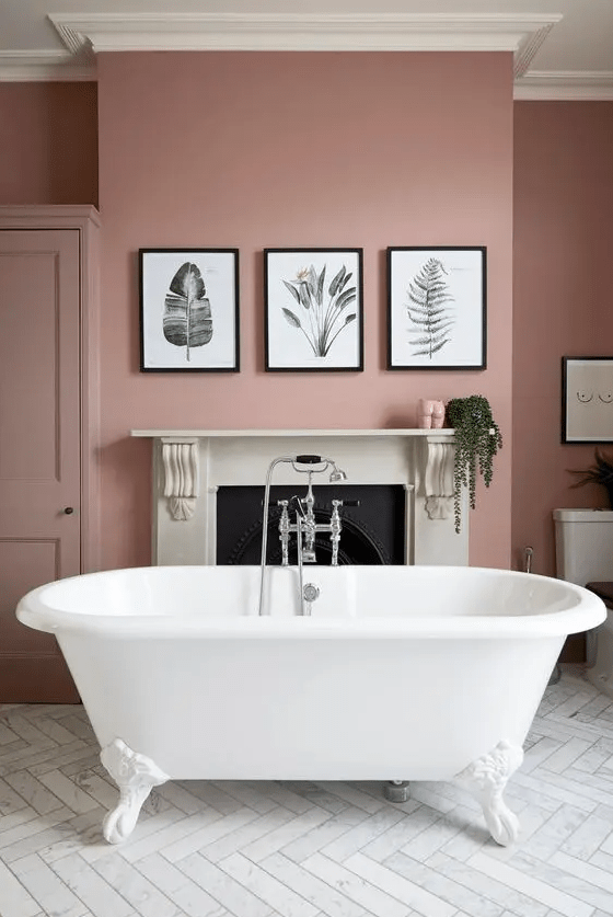 A chic pink bathroom with a non working fireplace, a white clawfoot tub, a gallery wall and some greenery