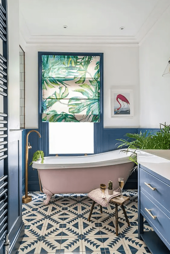 a lovely bathroom with blue paneling, a blue vanity, a navy and white tiled floor, a blush clawfoot tub and potted greenery and gold fixtures