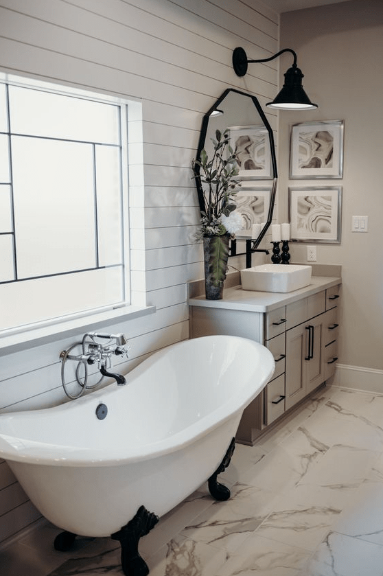 a modern farmhouse bathroom with white beadboard walls, a vintage tub, a wooden vanity and a black sconce