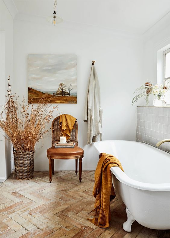 a serene bathroom with grey and terracotta tiles, a clawfoot tub, a vintage chair, mustard blankets and some branches