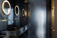 a stylish modern bathroom in black done with marble and stone, with lit up mirrors and brass touches