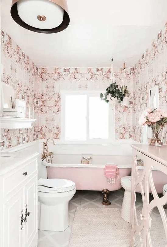 a stylish vintage-inspired bathroom done with pink printed wallpaper, a pink bathtub, a pink sideboard and brass fixtures and legs