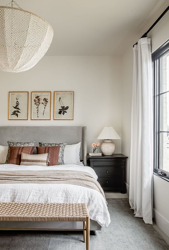 a lovely neutral bedroom with a grey bed and neutral bedding, a woven bench, a gallery wall, a black nightstand and some lamps