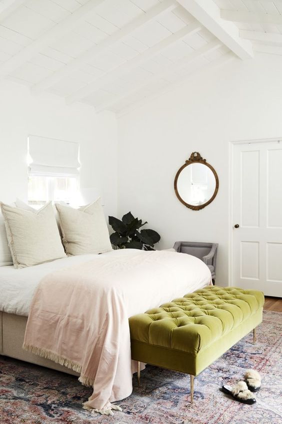 a modern bedroom with a bed and neutral bedding, a green upholstered bench, a boho rug and a grey chair, a mirror with a frame