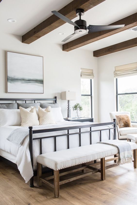 a modern bedroom with wooden beams, a bed and neutral bedding, a neutral upholstered bench, some artwork and a chair at the window