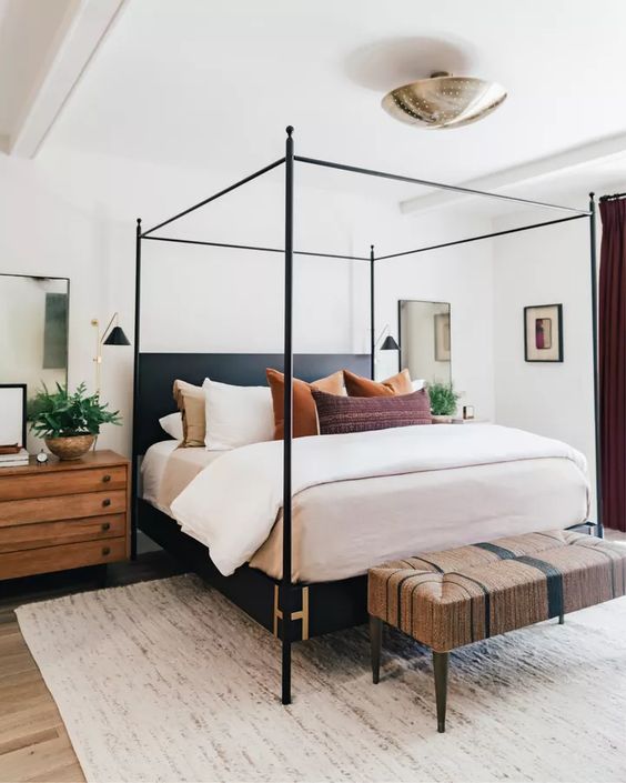 a modern farmhouse bedorom with a black frame bed and stained nightstands, a woven bench, some bold pillows and potted greenery