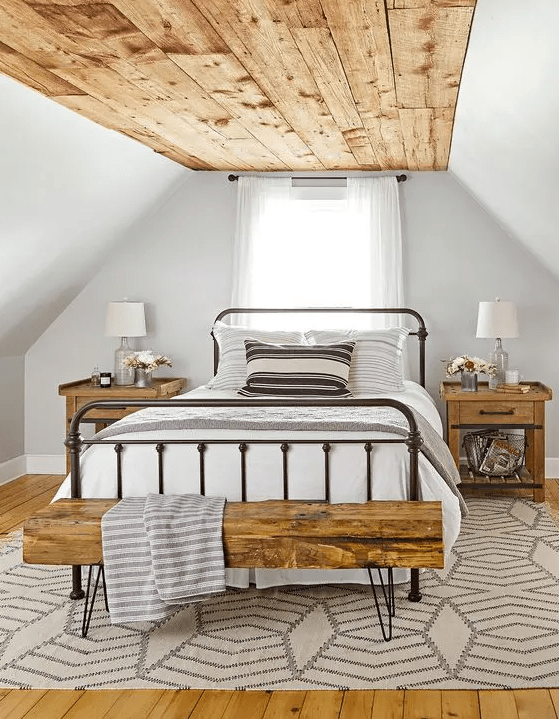 a modern farmhouse bedroom with a wooden ceiling, a wrought bed, wooden nightstands and a hairpin leg bench, some printed textiles