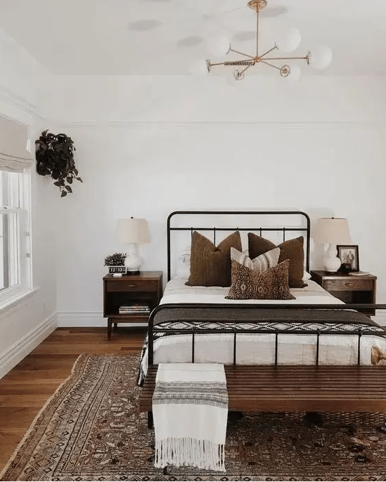 a modern farmhouse meets boho bedroom with a wrought bed and contrasting bedding, a stained bench, a printed rug, dark-stained nightstands and potted greenery