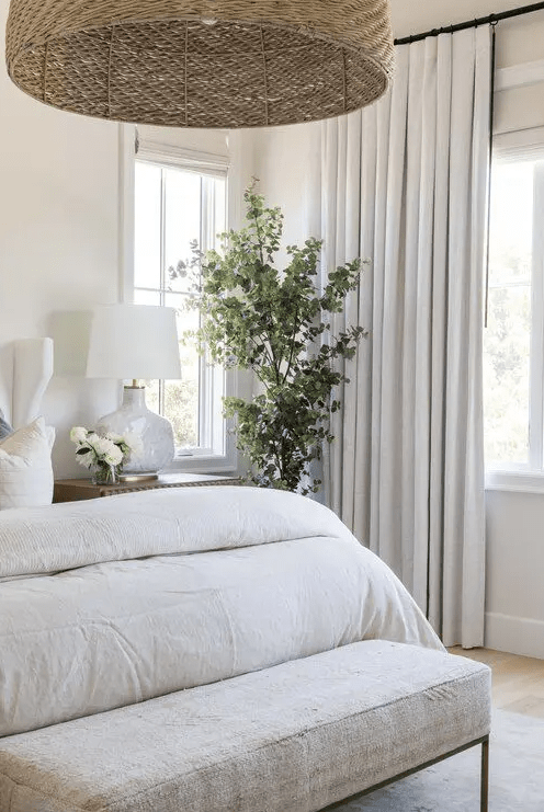 a neutral bedroom with a potted plant, neutral bedding and an upholstered bench, a woven pendant lamp