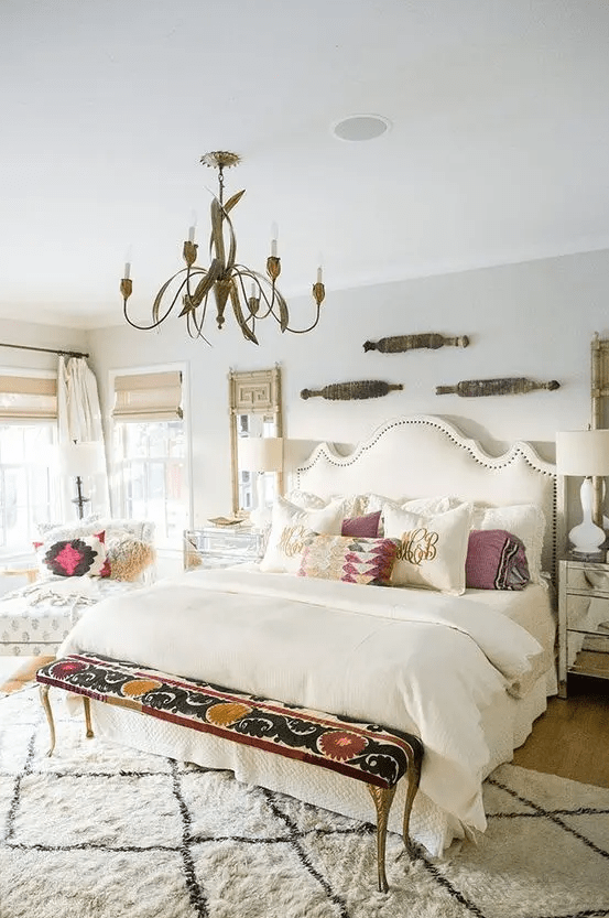 a neutral bedroom with an upholstered bed, printed pillows, a printed bench, a chair with printed pillows and a printed rug