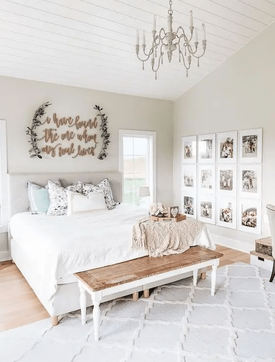 a neutral farmhouse space with an upholstered bed, a sign on the wall, a vintage chandelier, a wooden bench and a gallery wall