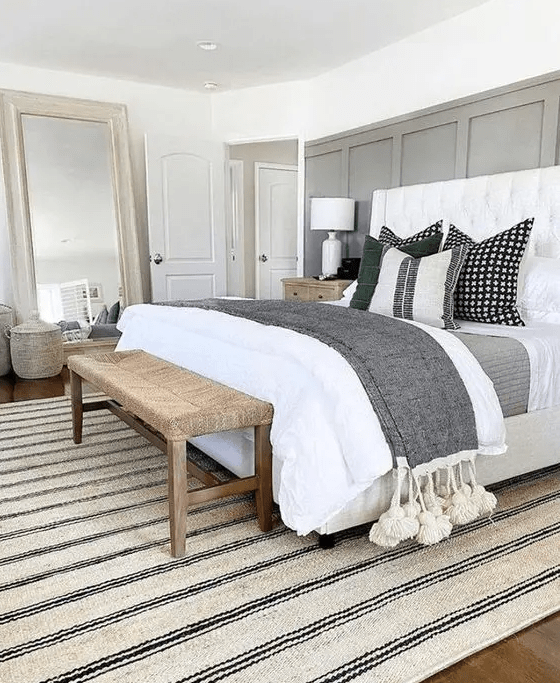 a neutral modern farmhouse bedroom with a grey paneled wall, a white bed with neutral bedding, a woven bench, a mirror and a rug