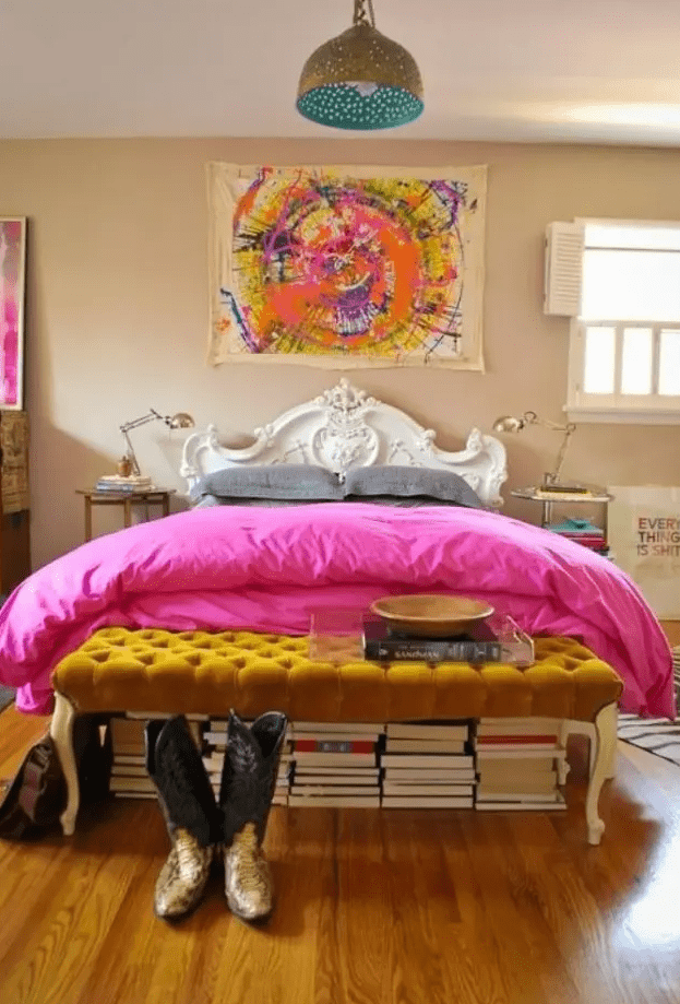 a super bright eclectic bedroom with bold artworks, a mustard tufted ottoman, stacks of books, a refined bed and mismatching nightstands