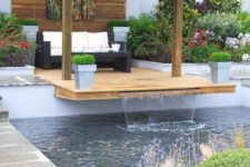 10 a pool cabana paired with a deck over the pool features a waterfall, potted plants and blooms, dark wicker furniture