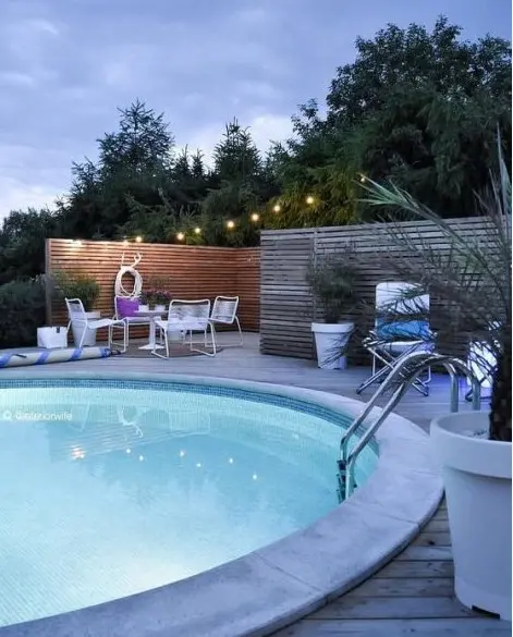 a beautiful stone deck with some outdoor furniture, a round lit up pool, some potted plants, lights and decor