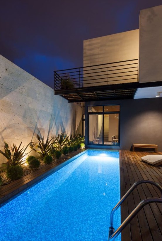 a bold outdoor tropical space with a long and narrow pool with lights, a wooden deck, growing plants and lights is cool