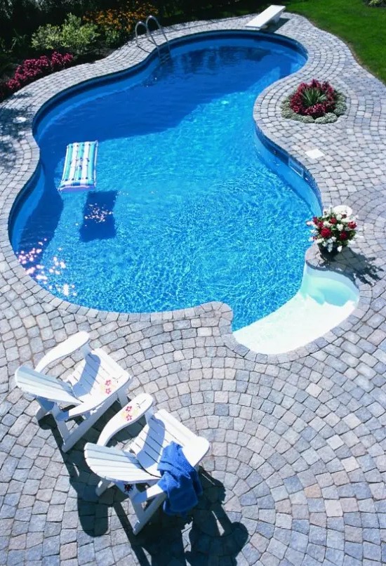 a catchily shaped pool with a whitewashed stone deck, simple white outdoor furniture, green lawn and bright blooms