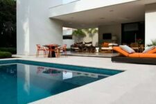 a contemporary outdoor space with a teal pool, a neutral stone tile deck, a metal table and orange chairs, some wicker furniture